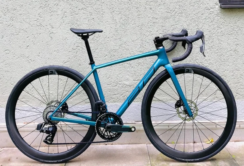 BQ Gravel, petrolfarbenes Carbon-Gravelbike, Seitenansicht vor einer Hauswand