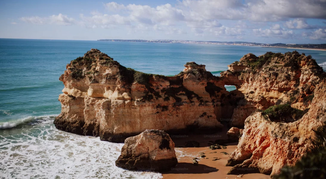 Goldgelbe Felsen mit einem Felsentor an einem Sandstrand der Algarve, davor Brandung, dahinter die Küste im Dunst.