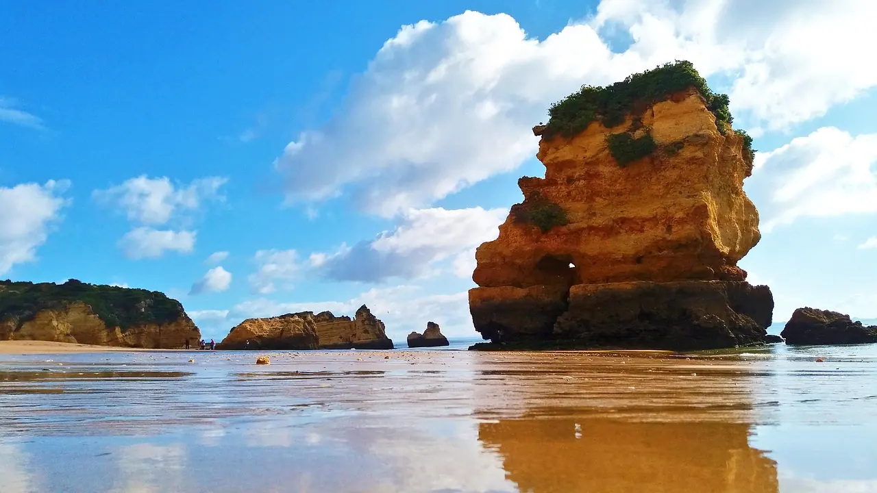 Goldgelber Felsen mit grünem Bewuchs am Sandstrand bei Lagos, davor spiegelt sich der Himmel im nassen Sand