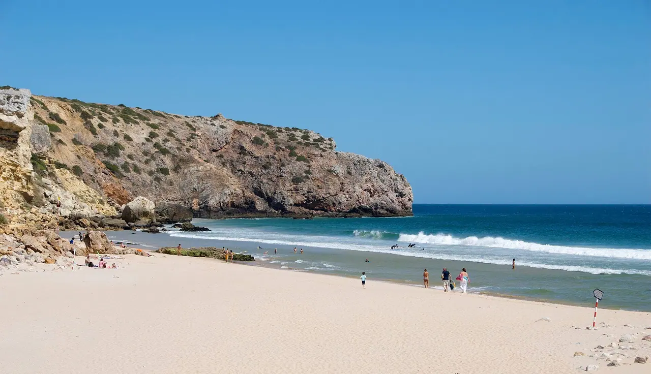 Heller Sandstrand der Praia do Zavial mit Brandung und einer steil abfallenden Felsküste im Hintergrund